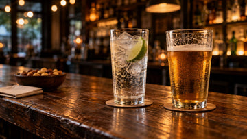 A pint of lager beside a tall glass of sparkling water with ice and lime on a worn wooden bar table, with warm string lights glowing in the background
