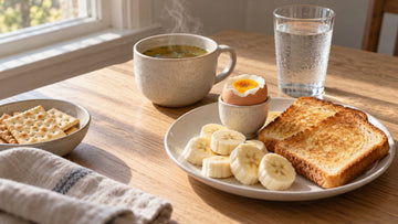 A plate of plain toast, sliced banana and a soft-boiled egg beside a mug of steaming broth, a glass of water and a bowl of crackers on a wooden table in morning light