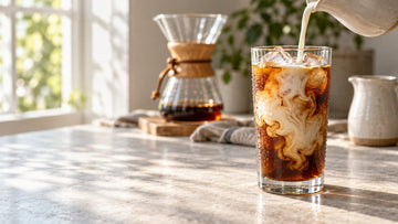 Cold milk being poured into a tall glass of iced coffee on a kitchen counter beside a glass pour-over coffee maker