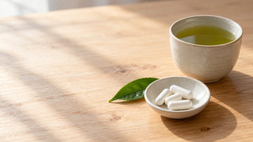 A cup of green tea on a table beside a small open dish of white supplement capsules and a fresh tea leaf in soft daylight