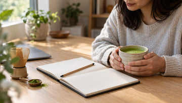 Hands holding a cup of whisked matcha at a wooden desk beside an open notebook, a bamboo whisk and a scoop of matcha powder