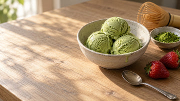 Scoops of pale green matcha ice cream in a ceramic bowl with a small dish of matcha powder and a bamboo whisk nearby