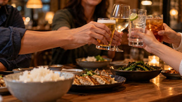 Friends toasting with beer, wine, sparkling water and iced tea over a dinner of dumplings, rice and greens